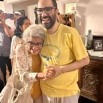 A photo of a British-Egyptian man hugging his elderly mother and smiling. He wears glasses and a dark beard with short hair and a yellow T-shirt. His mother has white hair and glasses and leans into his chest as they hold hands and smile