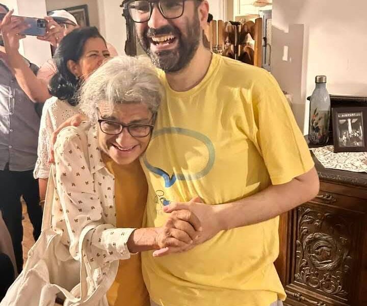 A photo of a British-Egyptian man hugging his elderly mother and smiling. He wears glasses and a dark beard with short hair and a yellow T-shirt. His mother has white hair and glasses and leans into his chest as they hold hands and smile