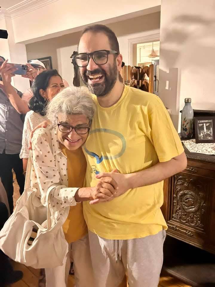 A photo of a British-Egyptian man hugging his elderly mother and smiling. He wears glasses and a dark beard with short hair and a yellow T-shirt. His mother has white hair and glasses and leans into his chest as they hold hands and smile